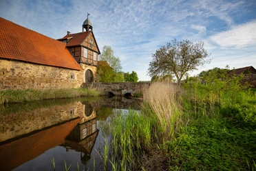 Historisches Fachwerkgebäude an einem Wassergraben mit Steinbrücke, von Vegetation umgeben.