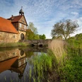Historisches Fachwerkgebäude an einem Wassergraben mit Steinbrücke, von Vegetation umgeben.