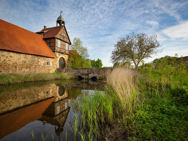 Historisches Fachwerkgebäude an einem Wassergraben mit Steinbrücke, von Vegetation umgeben.