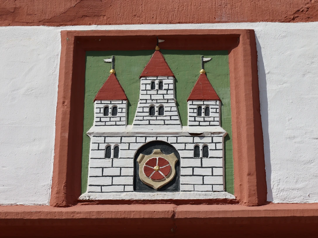 Wappenrelief mit drei Türmen und rotem Schild in einem Ziegelrahmen an einer weiß verputzten Wand.