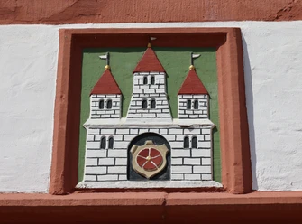 0W1A0206.jpg Wappenrelief mit drei Türmen und rotem Schild in einem Ziegelrahmen an einer weiß verputzten Wand.