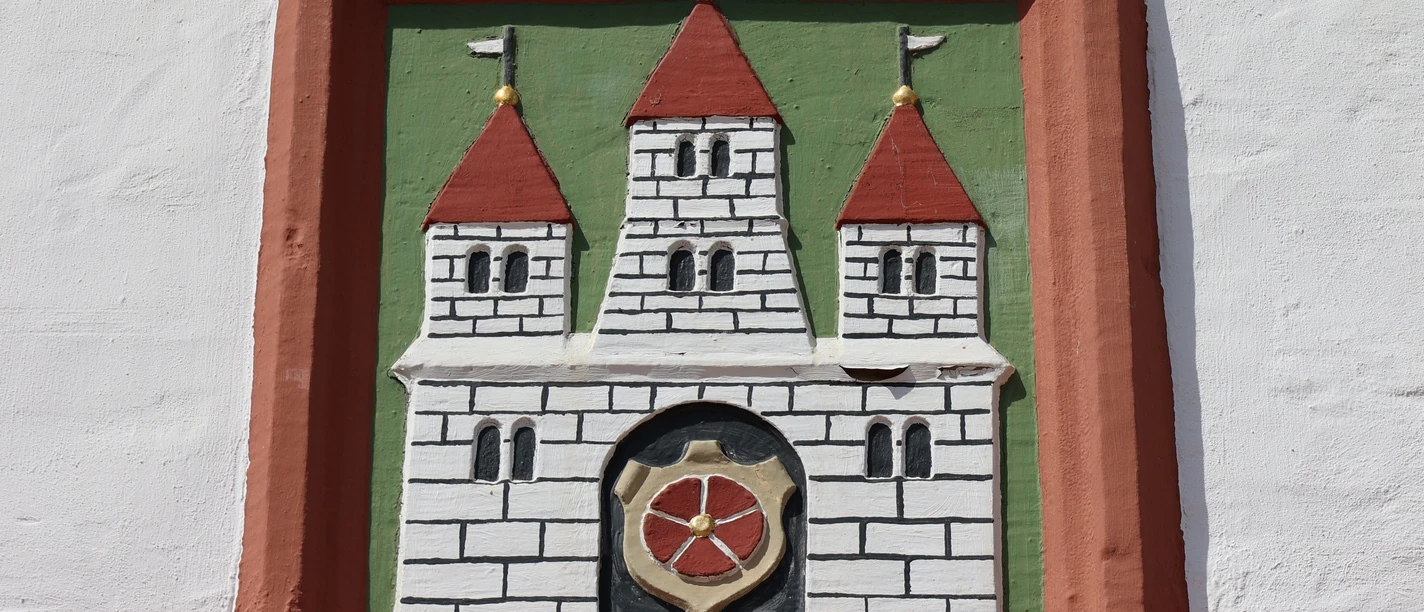 0W1A0206.jpg Wappenrelief mit drei Türmen und rotem Schild in einem Ziegelrahmen an einer weiß verputzten Wand.