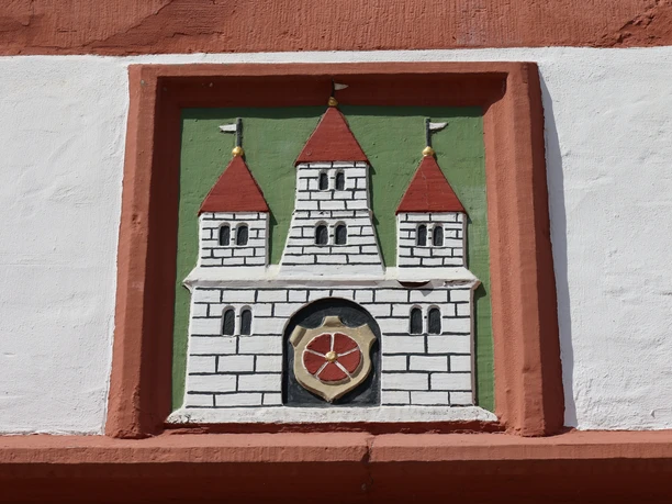 0W1A0206.jpg Wappenrelief mit drei Türmen und rotem Schild in einem Ziegelrahmen an einer weiß verputzten Wand.