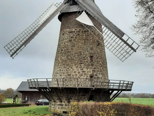 Eilhauser Königsmühle Historische Windmühle aus Stein in ländlicher Umgebung mit großen Holzflügeln, umgeben von Bäumen.