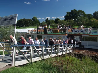 hx_weserdampfer_sigurdelert (3).JPG Menschen verlassen ein Ausflugsschiff in grüner Umgebung unter blauem Himmel, nahe Höxter.