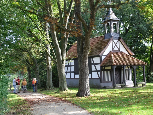 Rochuskapelle Eine kleine Fachwerkkapelle steht umgeben von Bäumen, mit Menschen seitlich auf einem Weg.