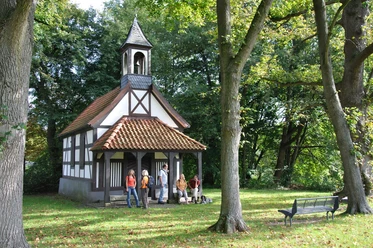 Kleine Fachwerkkapelle umgeben von Bäumen, mit Personen und einer Bank auf einer grünen Wiese.