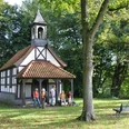 Kleine Fachwerkkapelle umgeben von Bäumen, mit Personen und einer Bank auf einer grünen Wiese.