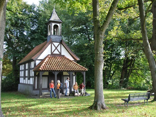 Rochuskapelle Kleine Fachwerkkapelle umgeben von Bäumen, mit Personen und einer Bank auf einer grünen Wiese.