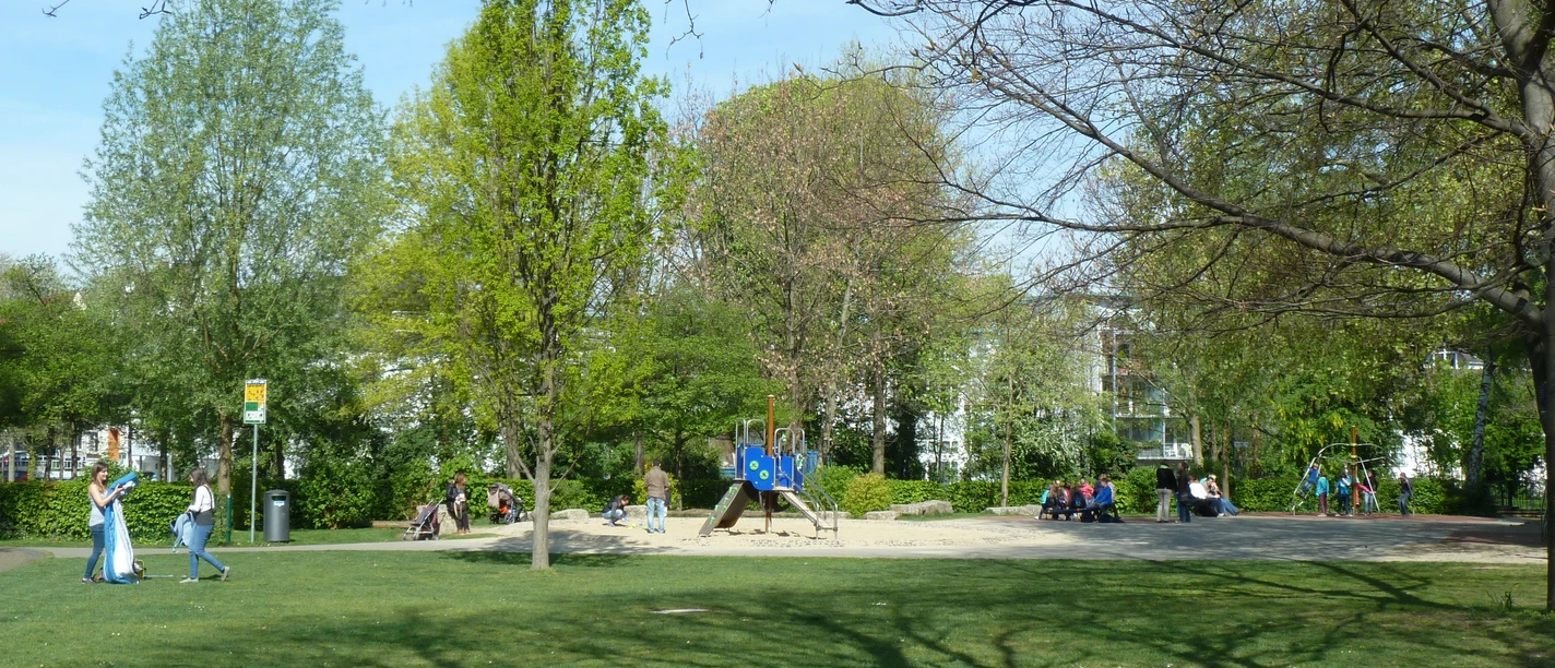 Spielplatz im Paderquellgebiet Grüner Spielplatz mit Klettergerüst und Sitzbereich zwischen Bäumen, Menschen genießen einen sonnigen Tag.