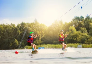 Zwei Kinder auf Wakeboards, gezogen von einem Seil über die Wasseroberfläche, unter strahlendem Sonnenschein.