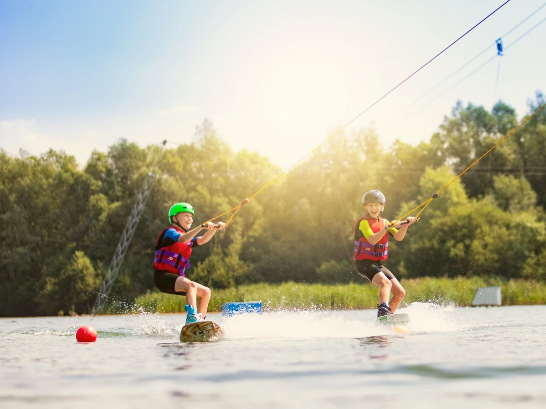 Zwei Kinder auf Wakeboards, gezogen von einem Seil über die Wasseroberfläche, unter strahlendem Sonnenschein.