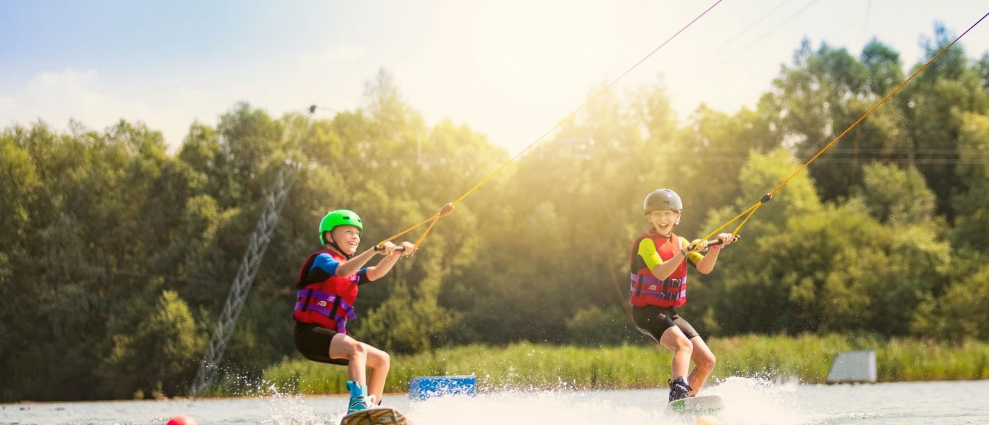 Zwei Kinder auf Wakeboards, gezogen von einem Seil über die Wasseroberfläche, unter strahlendem Sonnenschein.