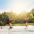 Wasserskianlage Nesthauser See Zwei Kinder auf Wakeboards, gezogen von einem Seil über die Wasseroberfläche, unter strahlendem Sonnenschein.