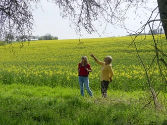 Zwei Frauen stehen vor einem blühenden Rapsfeld im Despental unter einem Baum.