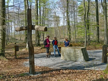 Drei Menschen stehen an einem Steintisch im Wald, umgeben von Bäumen und einem Holzkreuz.