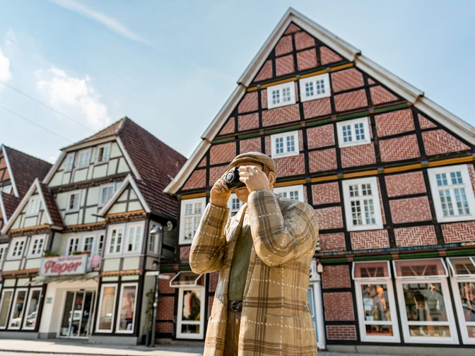 Eine Holzfigur in traditioneller Kleidung fotografiert Fachwerkhäuser in einer historischen deutschen Stadt.