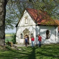 Eine kleine Kapelle mit rotem Ziegeldach steht in einer grünen, von Bäumen umgebenen Landschaft.