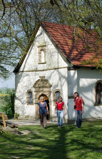 Eine kleine Kapelle mit rotem Ziegeldach steht in einer grünen, von Bäumen umgebenen Landschaft.