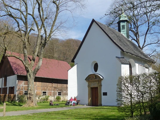 Kleine, weiße Kapelle neben einer großen Scheune, mehrere Personen sitzen im Grünen vor der Kapelle.