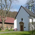 Kluskapelle St. Lucia Kleine, weiße Kapelle neben einer großen Scheune, mehrere Personen sitzen im Grünen vor der Kapelle.