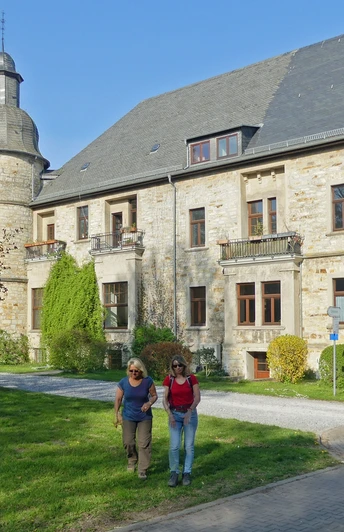Eine Gruppe von Menschen spaziert vor einer historischen Schlossgebäude mit Turm und Gartenanlage.