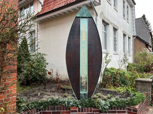 Metallene Stele mit der Aufschrift „STUWE BESTATTUNGEN“ umgeben von Pflanzen vor einem Haus.