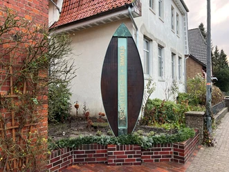Skulptur Das Portal Bücken Eine hohe, moderne Skulptur in Form eines tropfenförmigen Ovals neben einem roten Backsteingebäude.