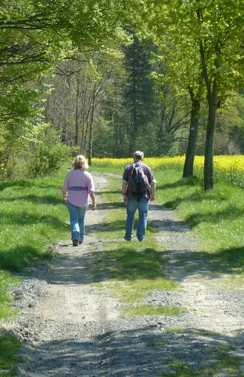 Zwei Menschen wandern im Frühling entlang eines von Bäumen gesäumten Weges, flankiert von Rapsfeldern.