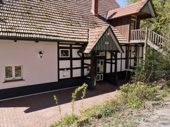 Fachwerkhaus mit grün-weißen Details, rotem Ziegeldach und Außenbereich, umgeben von Bäumen und Pflanzen.