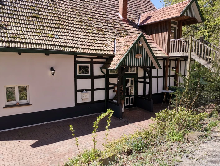 Café Waldkristall.jpg Fachwerkhaus mit grün-weißen Details, rotem Ziegeldach und Außenbereich, umgeben von Bäumen und Pflanzen.