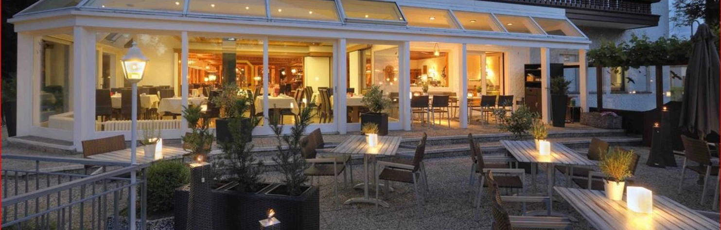 Deekes-Quellenhof_Luebbecke.JPG Restaurant-Bereich eines Gebäudes mit Außenterrasse, abends beleuchtet, mit Kerzen auf Tischen.