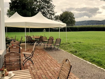 Kaffeeküche mit Zelt und einfachen Holztischen auf einer Terrasse neben einer grünen Wiese bei bewölktem Himmel.