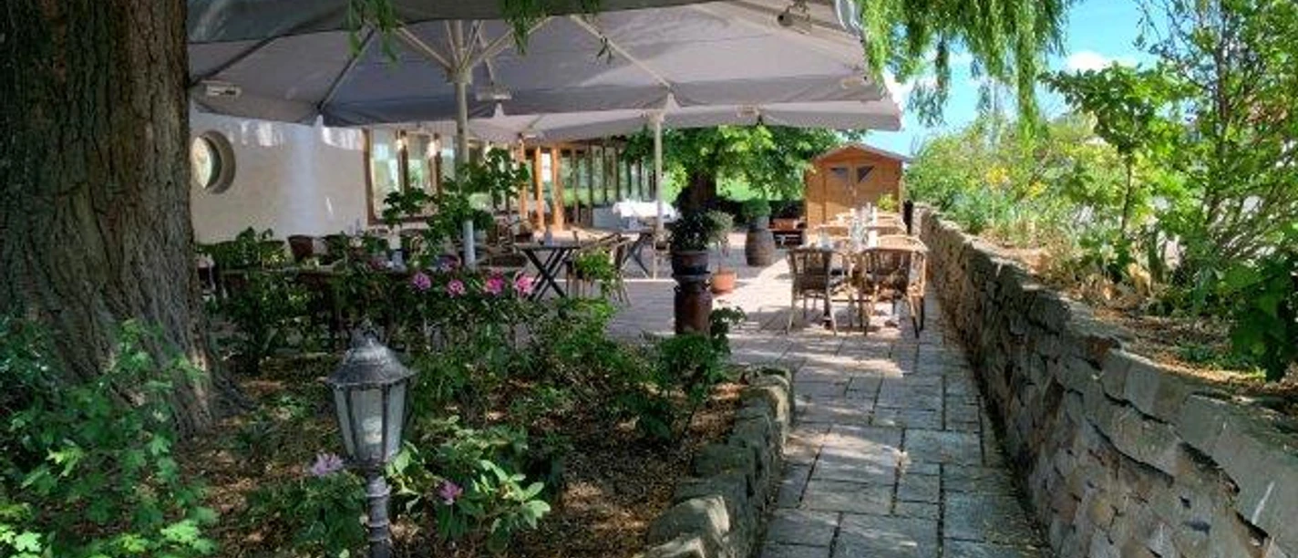 Dreimädelhaus Außenbereich eines Restaurants mit überdachten Sitzgelegenheiten, umgeben von grüner Natur und Backsteinmauer.