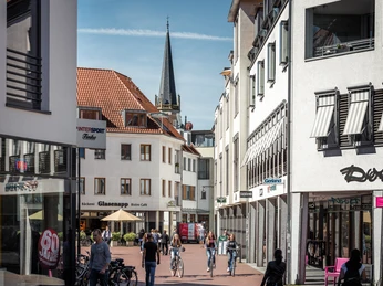 Kolbeplatz Gütersloh Eine belebte Fußgängerzone in einer Stadt mit Geschäften, Passanten und einem Kirchturm im Hintergrund.
