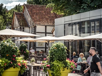 Gemütliche Cafés am Kolbeplatz Gütersloh Ein belebtes Café mit Sonnenschirmen und blühenden Pflanzen vor traditionellen Fachwerkhäusern.