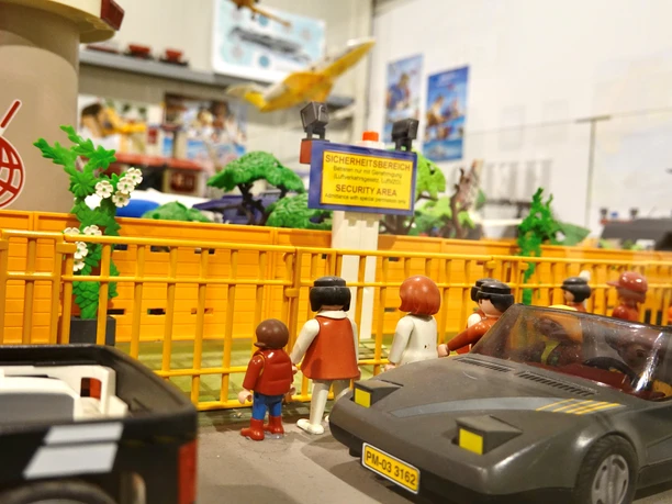 Miniwelten Lathen Modell einer Flughafen-Szene mit Spielfiguren, gelber Absperrung und Spielzeugautos in Miniaturform