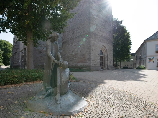 Kirchplatz in Steinheim Bronzestatue zweier Figuren vor einer historischen Kirche, umgeben von Bäumen und gepflastertem Platz.
