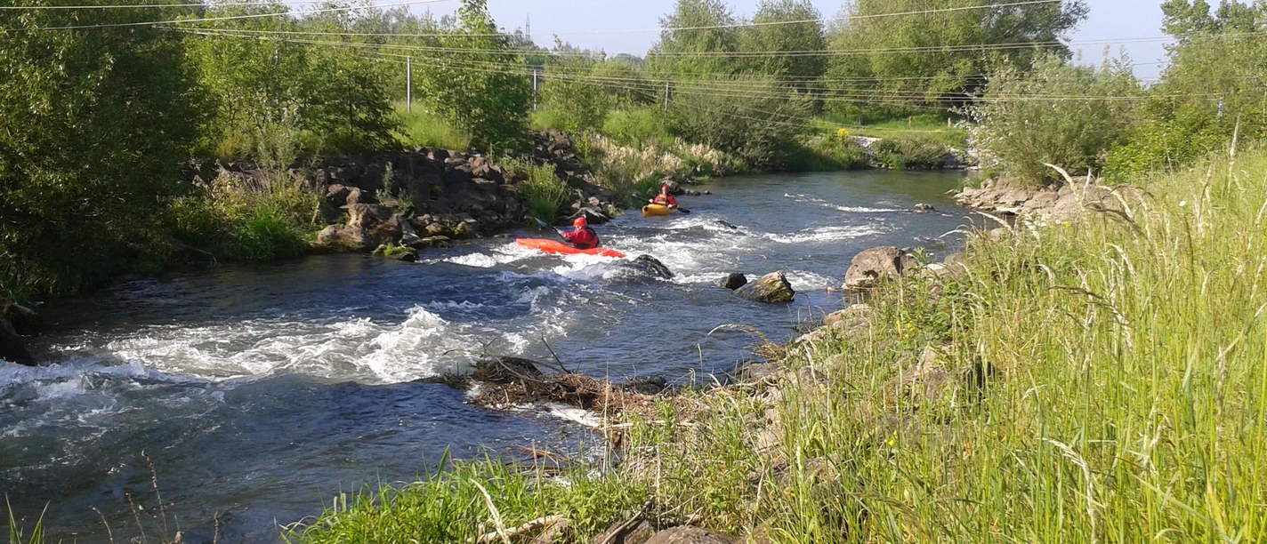Zwei Kanuten manövrieren ihre Boote durch das sprudelnde Wasser eines Flusses, umgeben von grüner Natur.