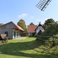 Historische Windmühle mit Fachwerkhäusern und Holzwagen in ländlicher Umgebung unter blauem Himmel.