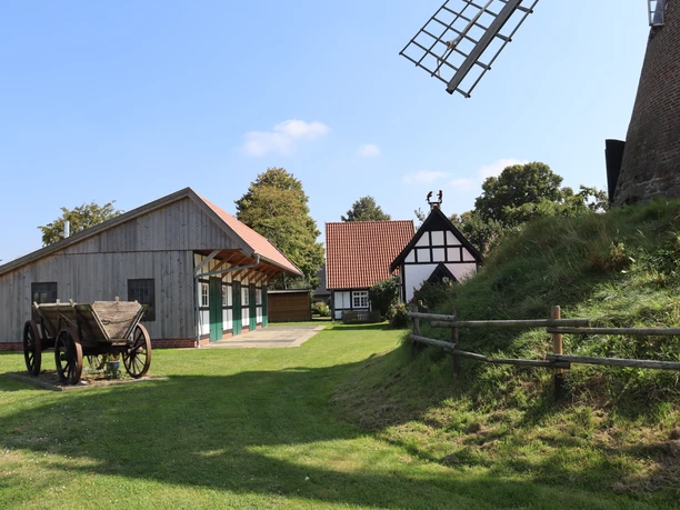Historische Windmühle mit Fachwerkhäusern und Holzwagen in ländlicher Umgebung unter blauem Himmel.
