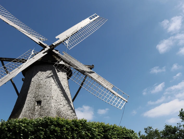 Historische Windmühle mit großen Flügeln, umgeben von grünem Laub, blauer Himmel im Hintergrund.