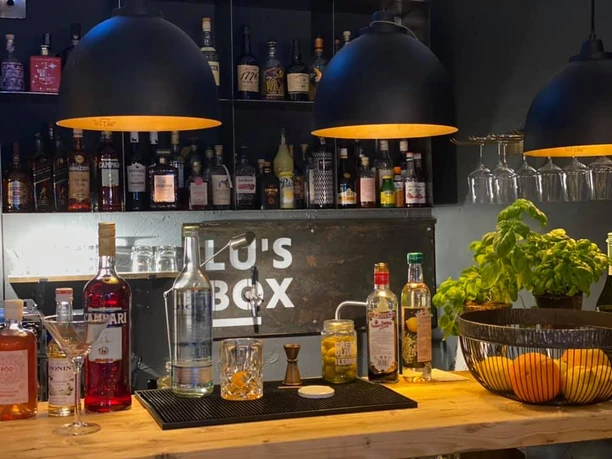 Lu´s Box Bar.jpg Gemütliche Bar mit Holztresen, verschiedenen Spirituosen und hängenden Gläsern beleuchtet durch Lampen.