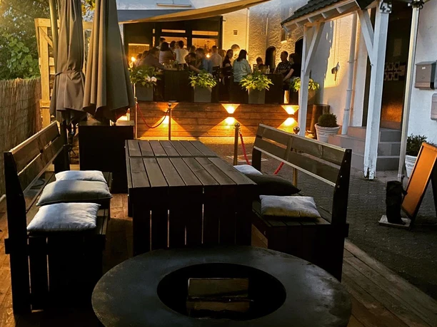 Lu´s Box Bistroterrasse.jpg Gemütliche Terrasse mit Holzmöbeln im Abendlicht, im Hintergrund eine gesellige, beleuchtete Runde.
