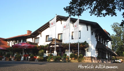 Gasthaus-Rose Gasthaus-Rose mit weißer Fassade und auffälligen Rosenaufhängungen, umgeben von hohen Fahnenmasten.
