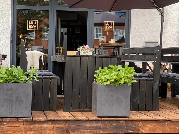 Eine gemütliche Caféterrasse mit Holzmöbeln, Pflanzkübeln und Sonnenschirm, Lu's Box im Hintergrund.
