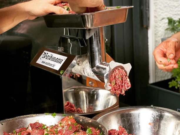 Frisches Hackfleisch wird von mehreren Personen durch einen Fleischwolf gedreht und in Schalen gesammelt.