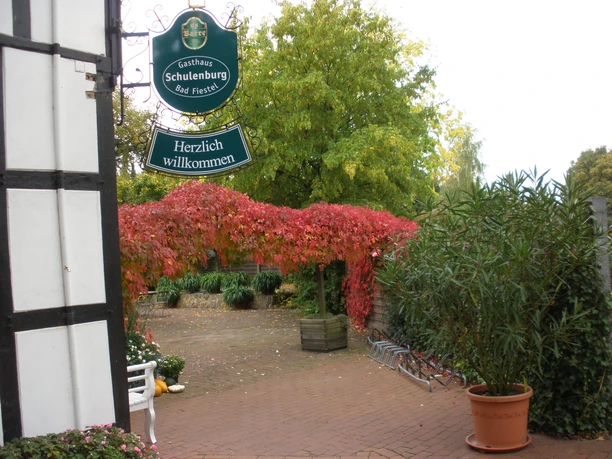 Gasthaus Schulenburg Eingang eines Gasthauses mit Schild, herbstlichem Laub, Fahrradständer und blühenden Pflanzen.