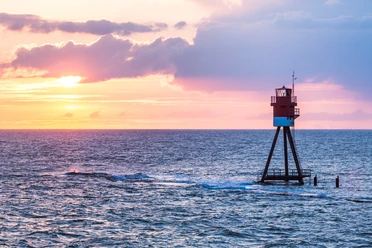 Fischerbalje Ein rot-weiß gestreifter Leuchtturm steht in der Weite des blauen Meeres bei Sonnenuntergang.