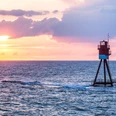 Fischerbalje Ein rot-weiß gestreifter Leuchtturm steht in der Weite des blauen Meeres bei Sonnenuntergang.